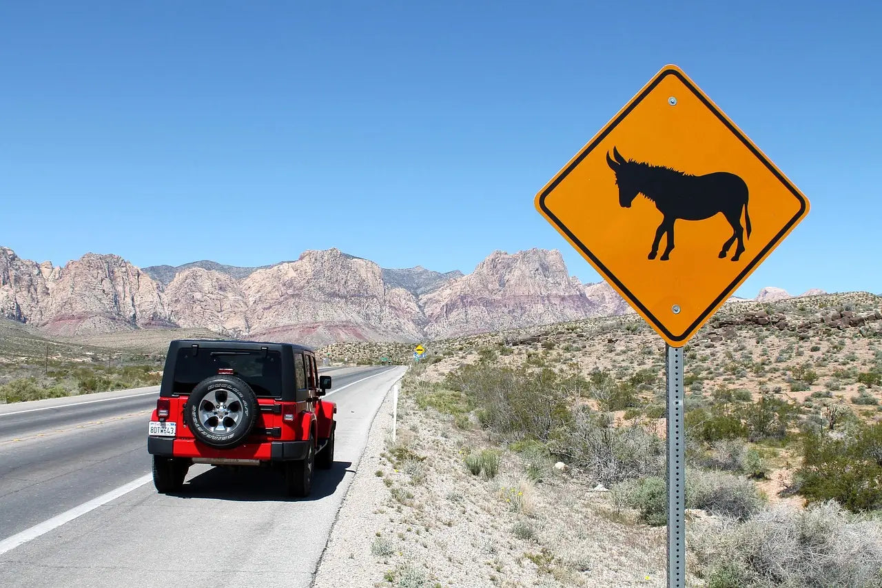 A red jeep wrangler no doors configuration parked along a scenic desert highway during an open-air road trip, featuring a wild donkey warning sign and rugged mountain landscapes.