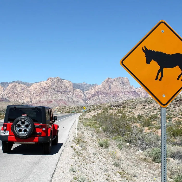 A red jeep wrangler no doors configuration parked along a scenic desert highway during an open-air road trip, featuring a wild donkey warning sign and rugged mountain landscapes.