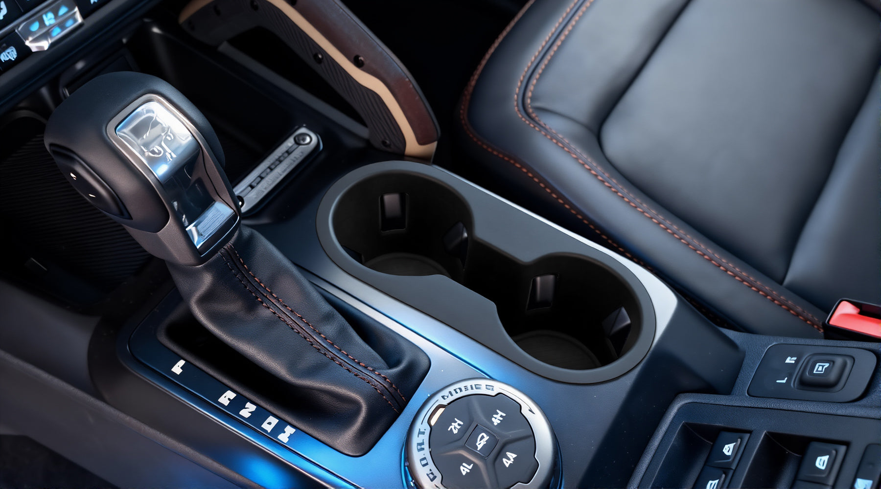 Close-up of a car center console featuring an automatic gear shifter, cup holders, drive mode selector, and black leather seats with orange stitching.